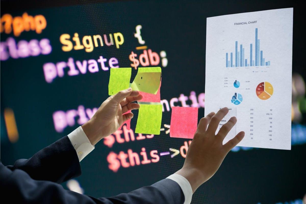 Person holding a financial chart with graphs and sticky notes against a background of colorful coding text on a screen.